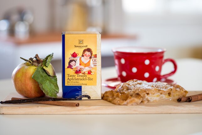 Foto einer Packung Tante Trudls Apfelstrudel-Tee. Daneben sieht man frisch gebackenem Apfelstrudel. | © SONNENTOR