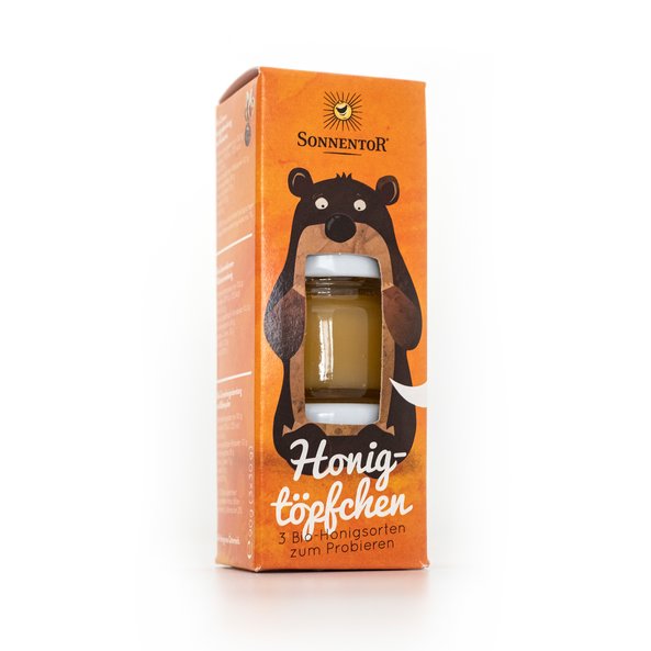 Eine orangefarbige Packung mit einem Bären darauf. Die Verpackung hat ein offenes Fenster durch das man den Honig sieht.