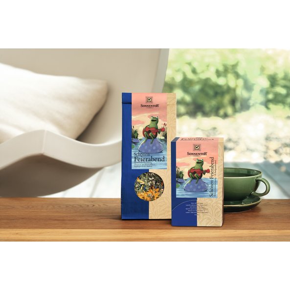 Foto einer Packung Schönen Feierabend Kräutertee lose, einer Packung Schönen Feierabend Kräutertee Doppelkammer und rechts davon eine dunkelgrüne Tasse Tee.