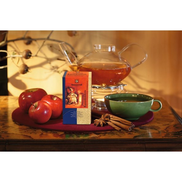 Foto von einer Packung Kaminknistern Früchtetee, einer Teekanne, einer dunkelgrünen Tasse Tee, drei rote Äpfel, zwei Sternanis und vier Zimtstangen.