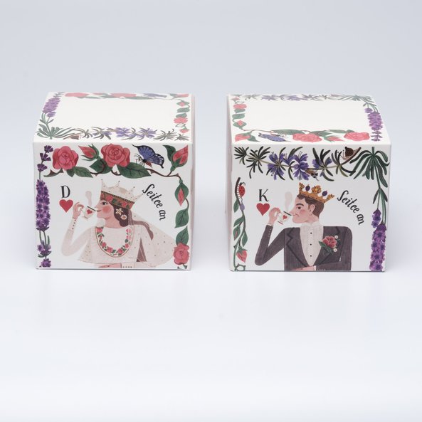 Auf dem Bild sind zwei Tee Schachteln zu sehen. Nebeneinander stehen die Herzkönigin Schachtel und die Herzkönig Schachtel. Beide verziert mit Blumen.