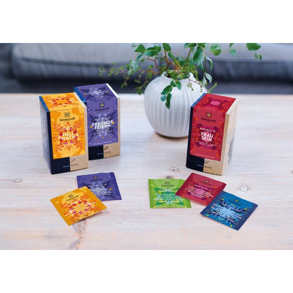 In the picture you can see three Happiness Is tea packs in the colors purple, red and yellow. These are standing on a table. Behind them you can see a plant.