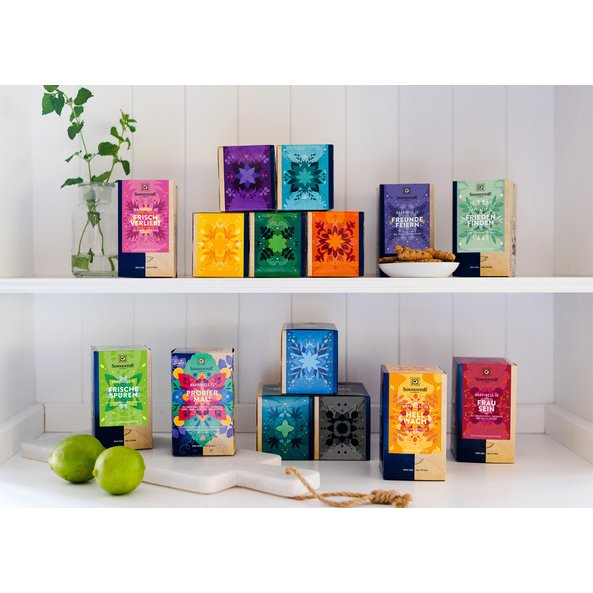 Auf dem Bild sind Teepackungen der Happiness Is Reihe zu sehen. Sie stehen in einem Regal und sind auf zwei Etagen verteilt.