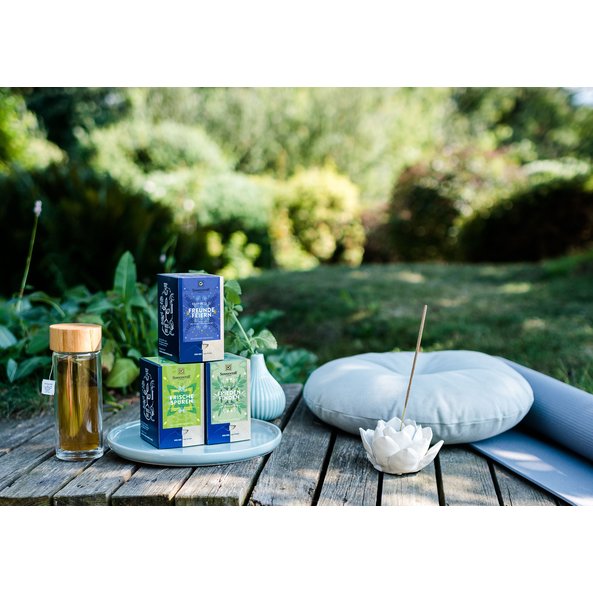 Auf dem Bild sieht man drei Happiness Is Teepackungen in der Natur stehen. Daneben steht eine Flasche Tee, eine Yogamatte und ein Kissen.