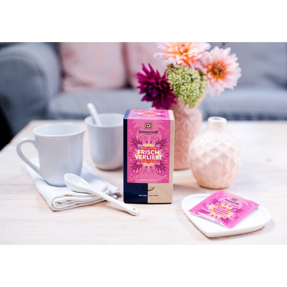 Zu sehen ist eine rosa Happiness Is Teepackung auf einem Tisch. Daneben stehen Teetassen und Blumen.