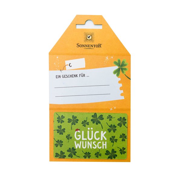 Auf dem Foto ist eine grüne Geschenkkarte mit dunkelgrünen Kleeblättern zu sehen. In der Mitte der Karte steht Glückwunsch. Im Hintergrund ist die gelbe Verpackung.