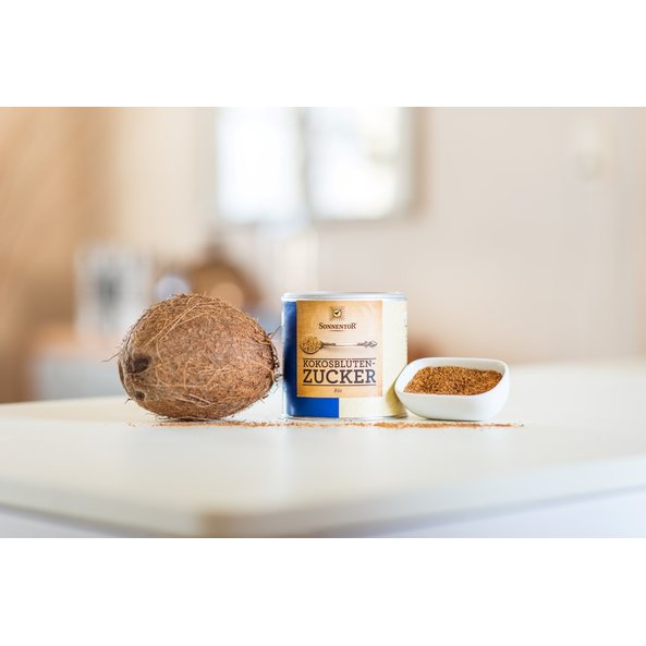 The photo shows a coconut and a jumbo spice tin coconut sugar. Next to them there is a small bowl filled with coconut sugar placed.