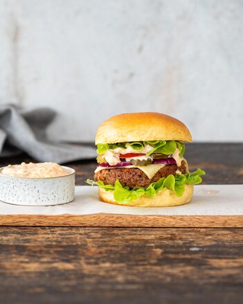 Der Cheeseburger ist gefüllt mit einem Rindfleischpattie, Salat, Paradeiser, Sauce, roten Zwiebeln und Käse und liegt auf einem Brett neben einem Schüsserl mit Sauce. | © SONNENTOR