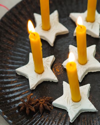 Auf dem Foto sieht man mehrere sternförmige Kerzenhalter aus Ton. Darin stecken orange färbige Kerzen die brennen. | © SONNENTOR