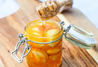 The garlic cloves are already in the jar with honey, and more honey is added from a honey spoon. The glass stands on a wooden board. | © SONNENTOR