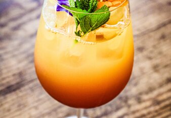 Der orangefarbene Cocktail ist in ein schönes Weinglas gefüllt, unten ist er dunkler als oben. Es befinden sich Eiswürfel mit im Glas und alles ist mit essbaren Blüten und Minzblättern dekoriert. | © SONNENTOR